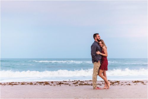 Girl in red dress snuggling up to finance on beach during Floridana Beach engagement