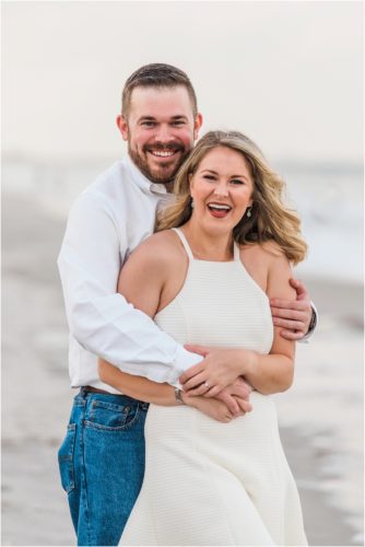 Man and girl in white dress hugging and laughing on beach during Surprise Proposal