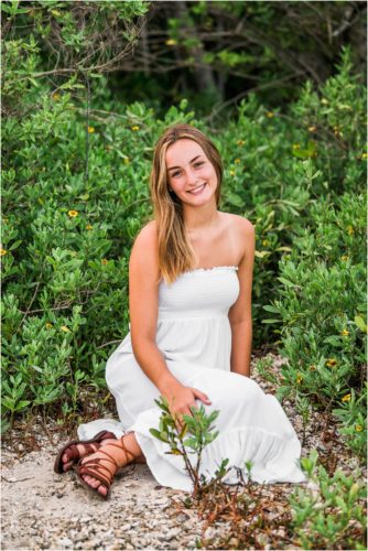 Girl in white dress sitting on beach with trees behind her