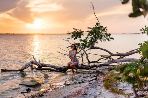Girl sitting on driftwood with sun setting in background