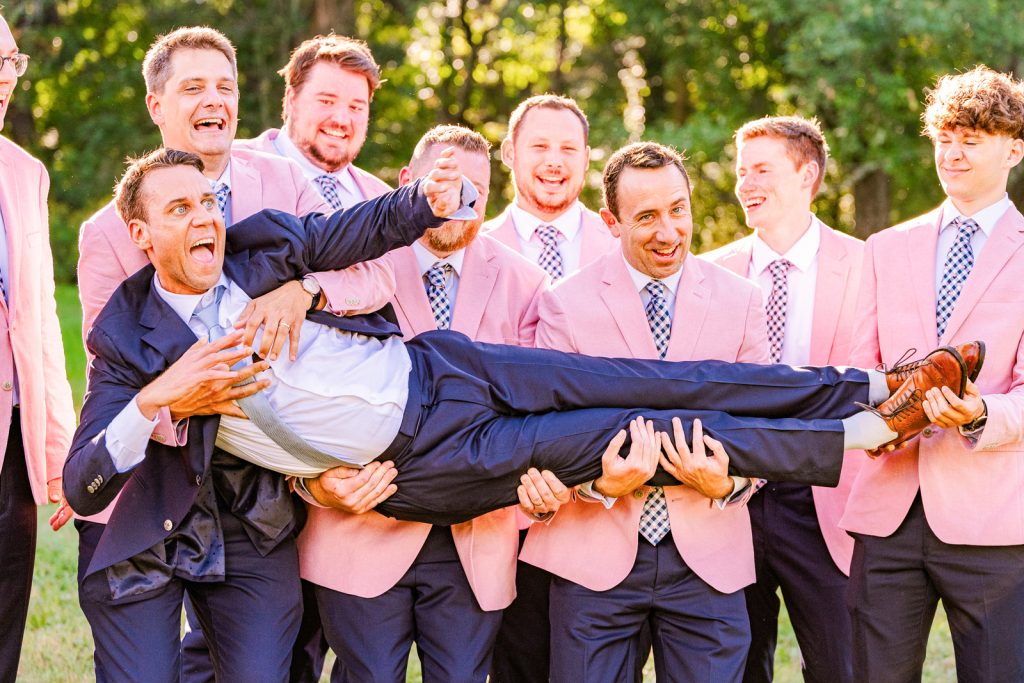 Groomsmen in pink jackets lifting up the groom and laughing at a wedding in Pawling New York
