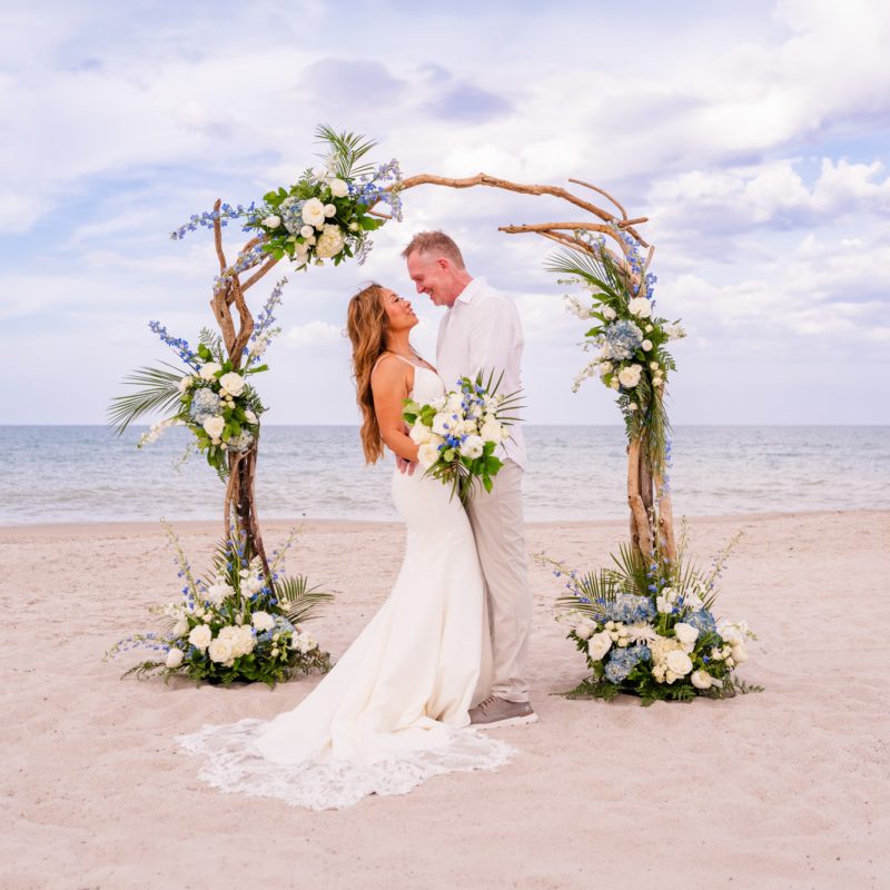 Bride and groom embracing under a floral driftwood arch on Melbourne beach