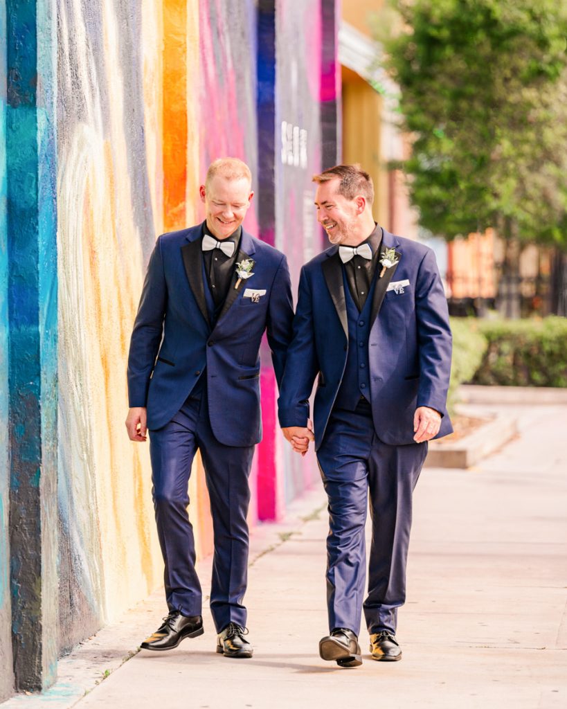 Two grooms walking together beside a colorful mural on their wedding day at Hotel Melby