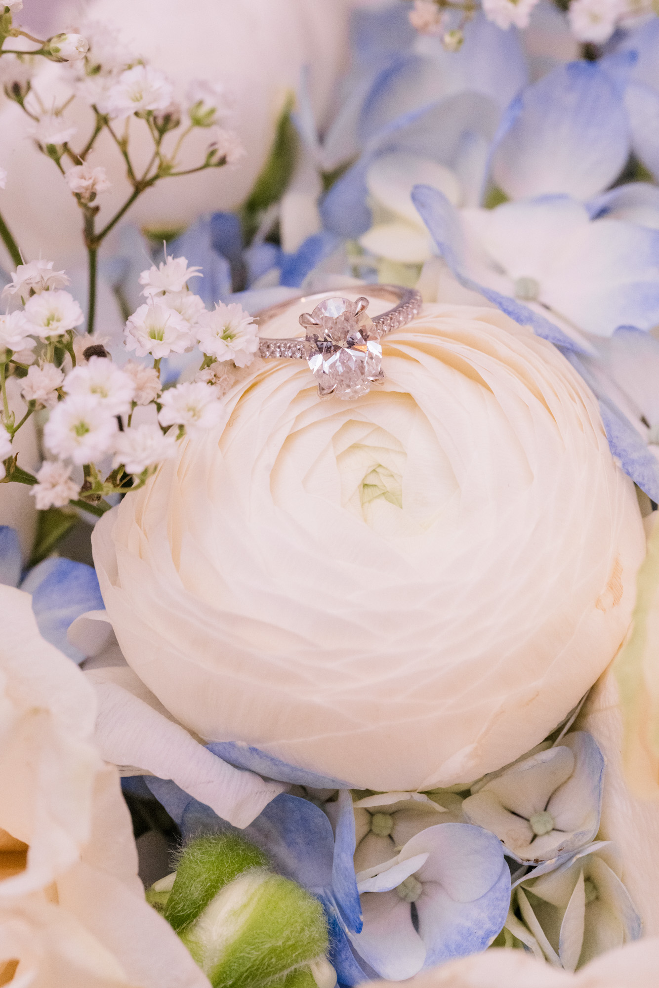 Diamond engagement ring resting on a white flower surrounded by soft blue wedding florals.