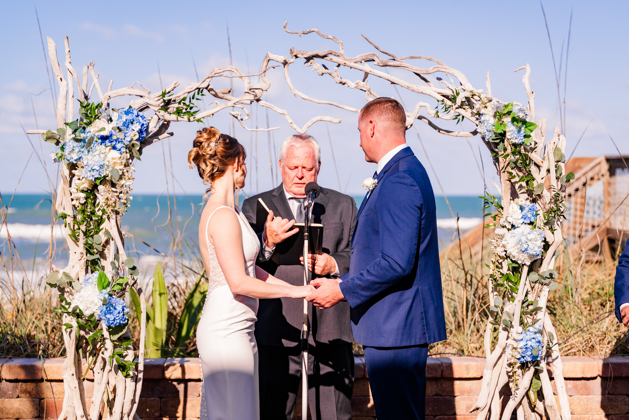 Bride and groom holding hands beneath a driftwood wedding arch overlooking the ocean.