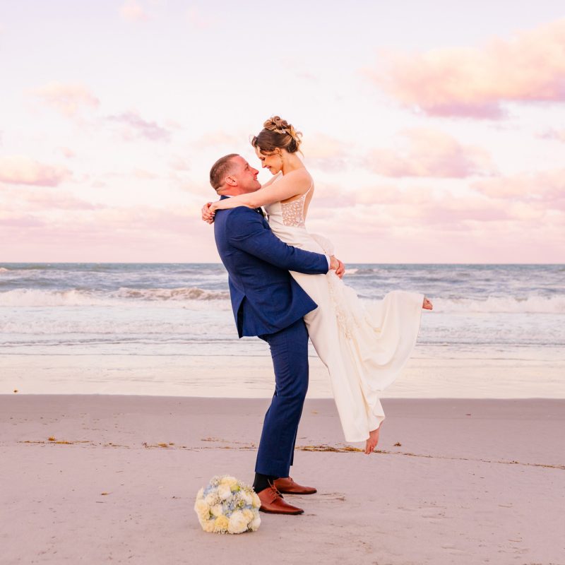 Groom lifting the bride on the beach beneath a pastel sunset sky.