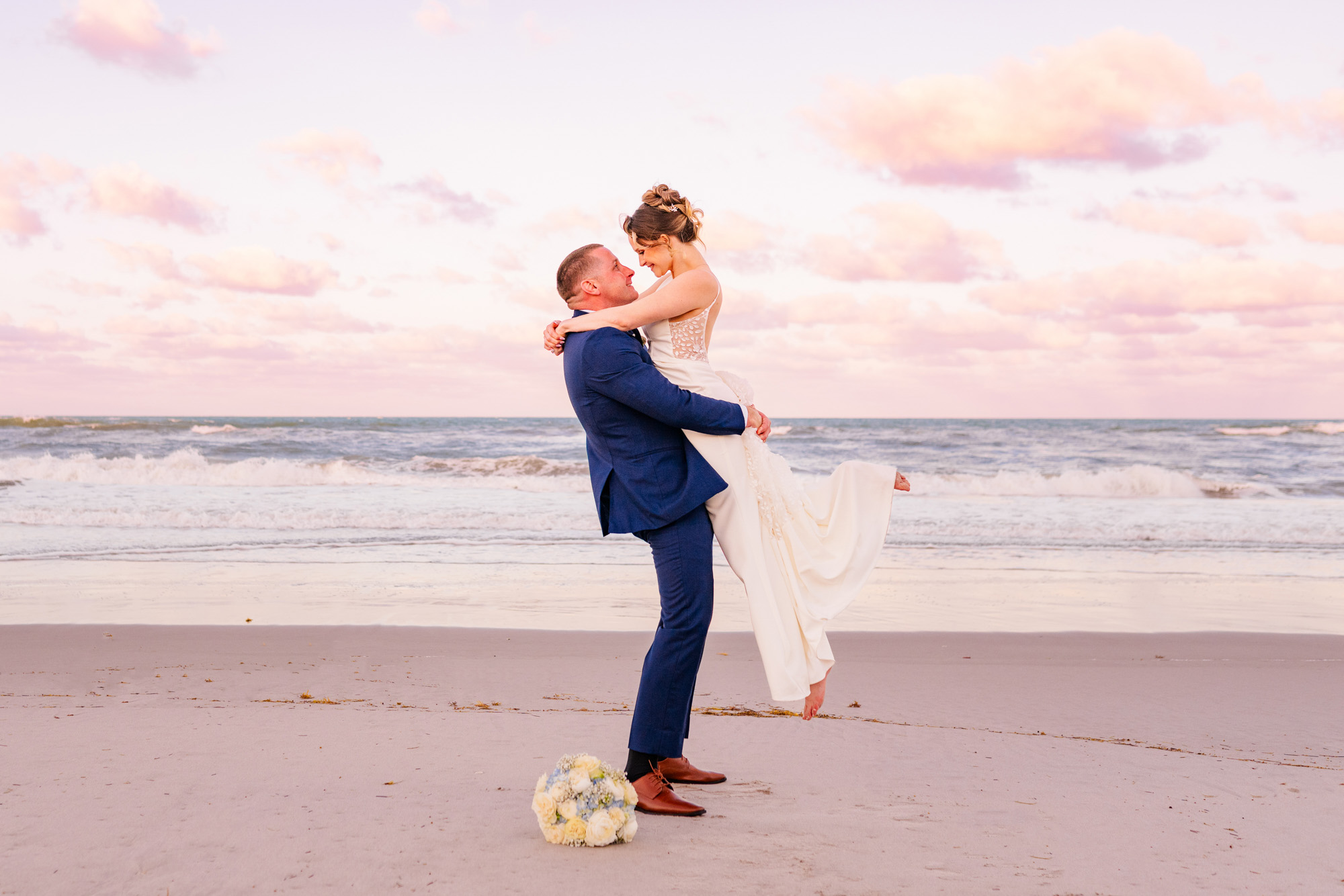 Groom lifting the bride on the beach beneath a pastel sunset sky.