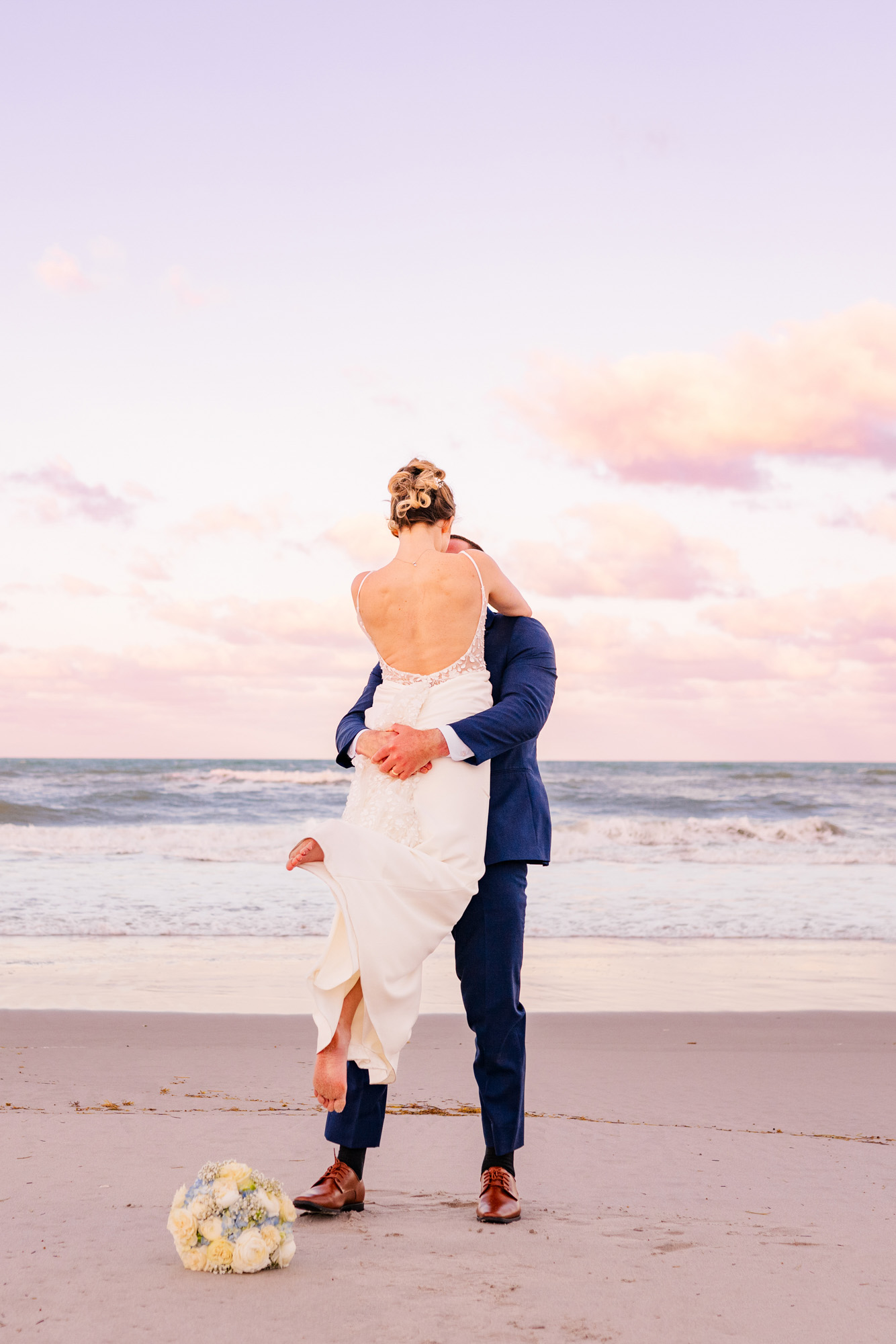 Bride and groom laughing together while he lifts her on the beach at sunset.