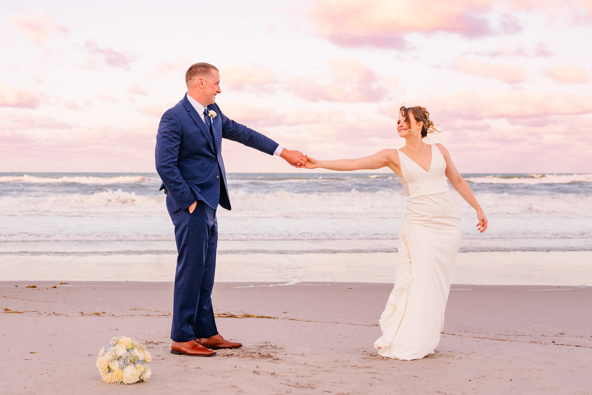 Bride and groom reaching toward one another during playful sunset beach portraits.