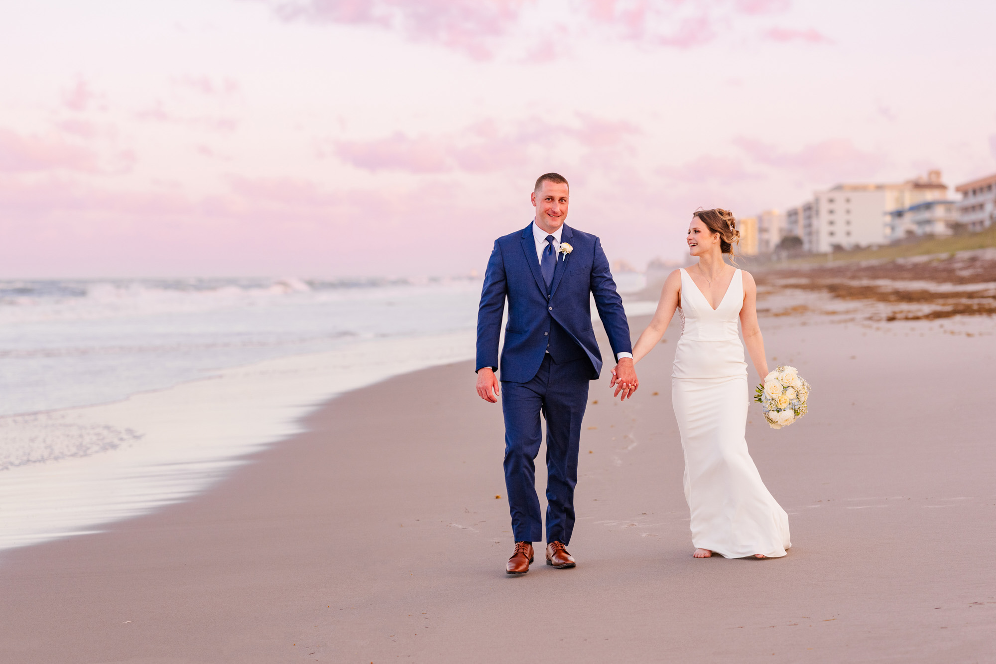 Bride and groom holding hands on the beach beneath soft pink clouds after their Harbour House Oceanfront wedding.