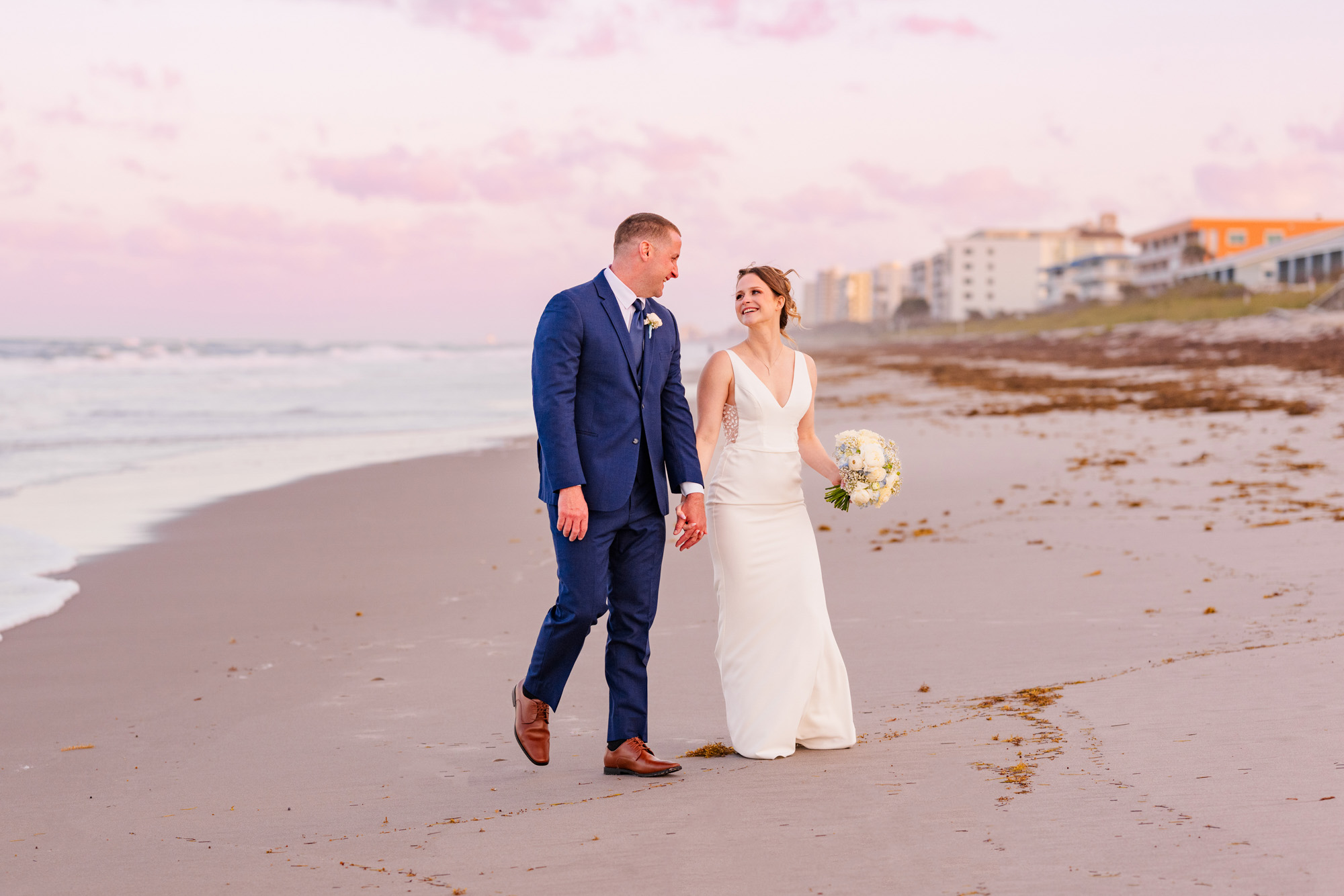 Bride and groom walking hand in hand along the beach beneath a pink sunset sky.