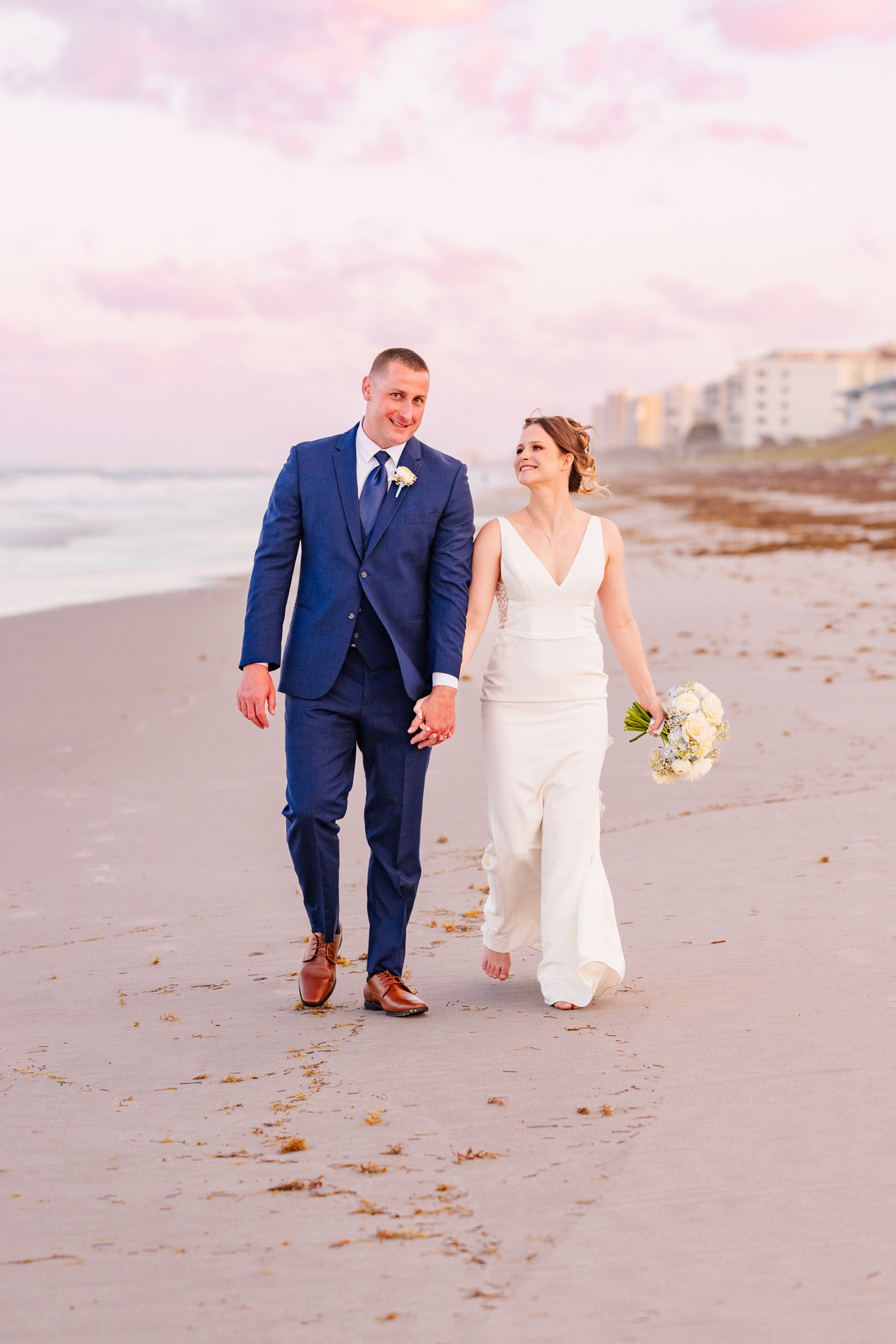 Bride and groom walking barefoot along the shoreline at sunset.