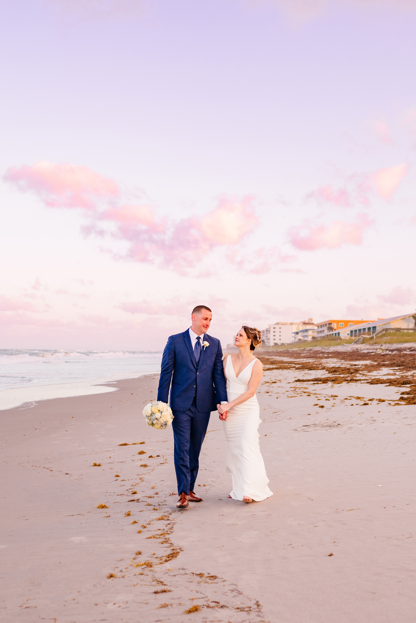 Newlyweds walking together on the beach beneath soft pink clouds.