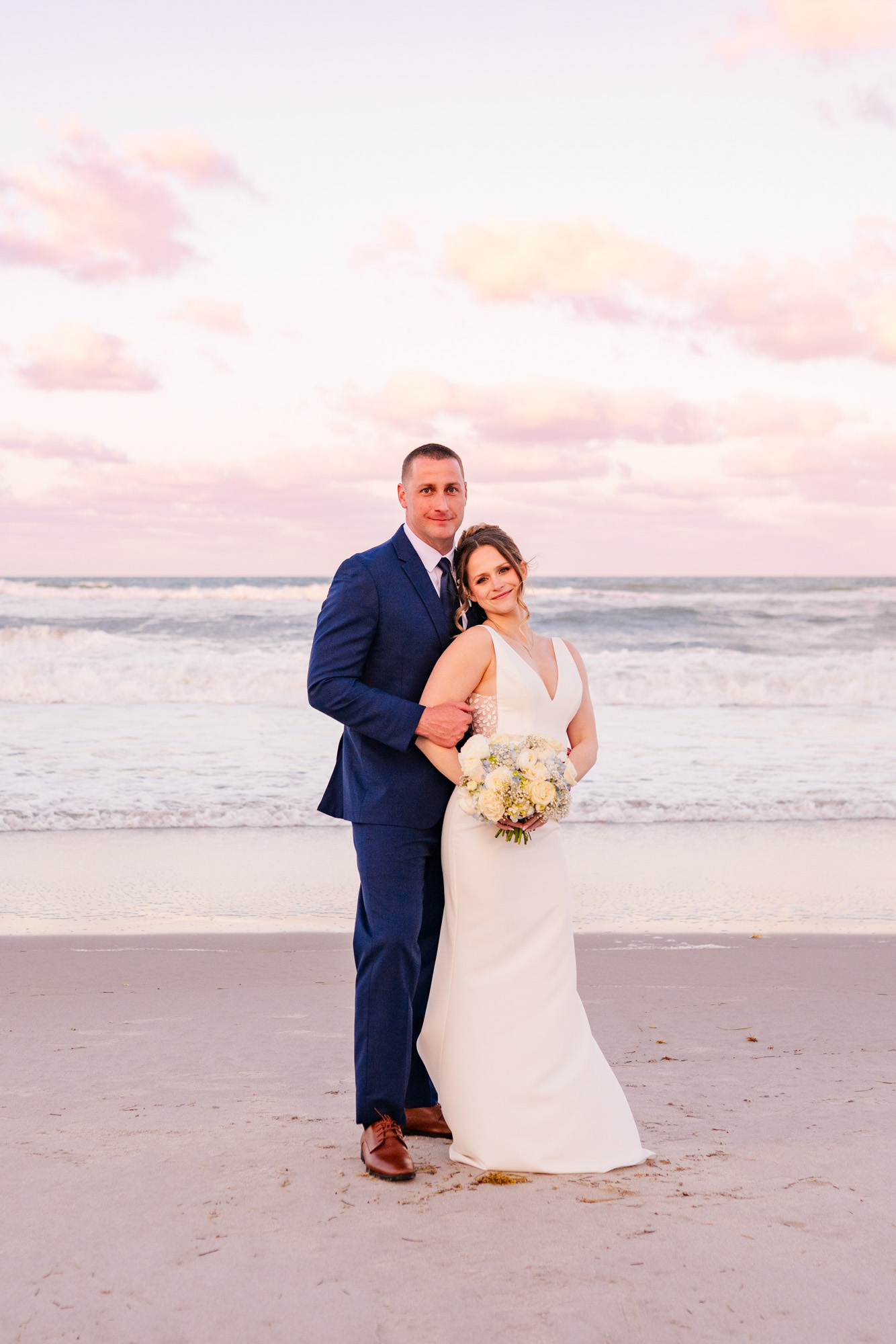 Newlyweds posing together on the beach with ocean waves and pastel clouds behind them.