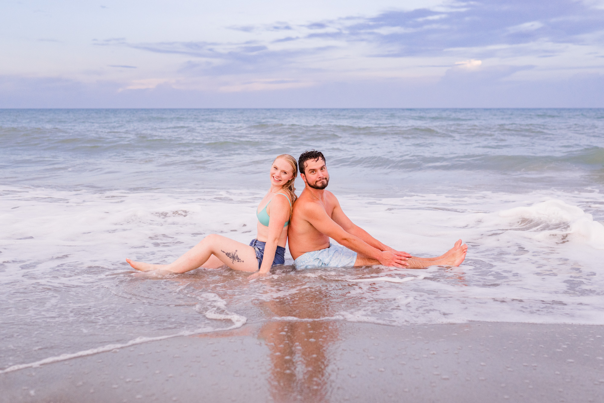 couple sitting back to back by the edge of the ocean with a wave about to crash over them