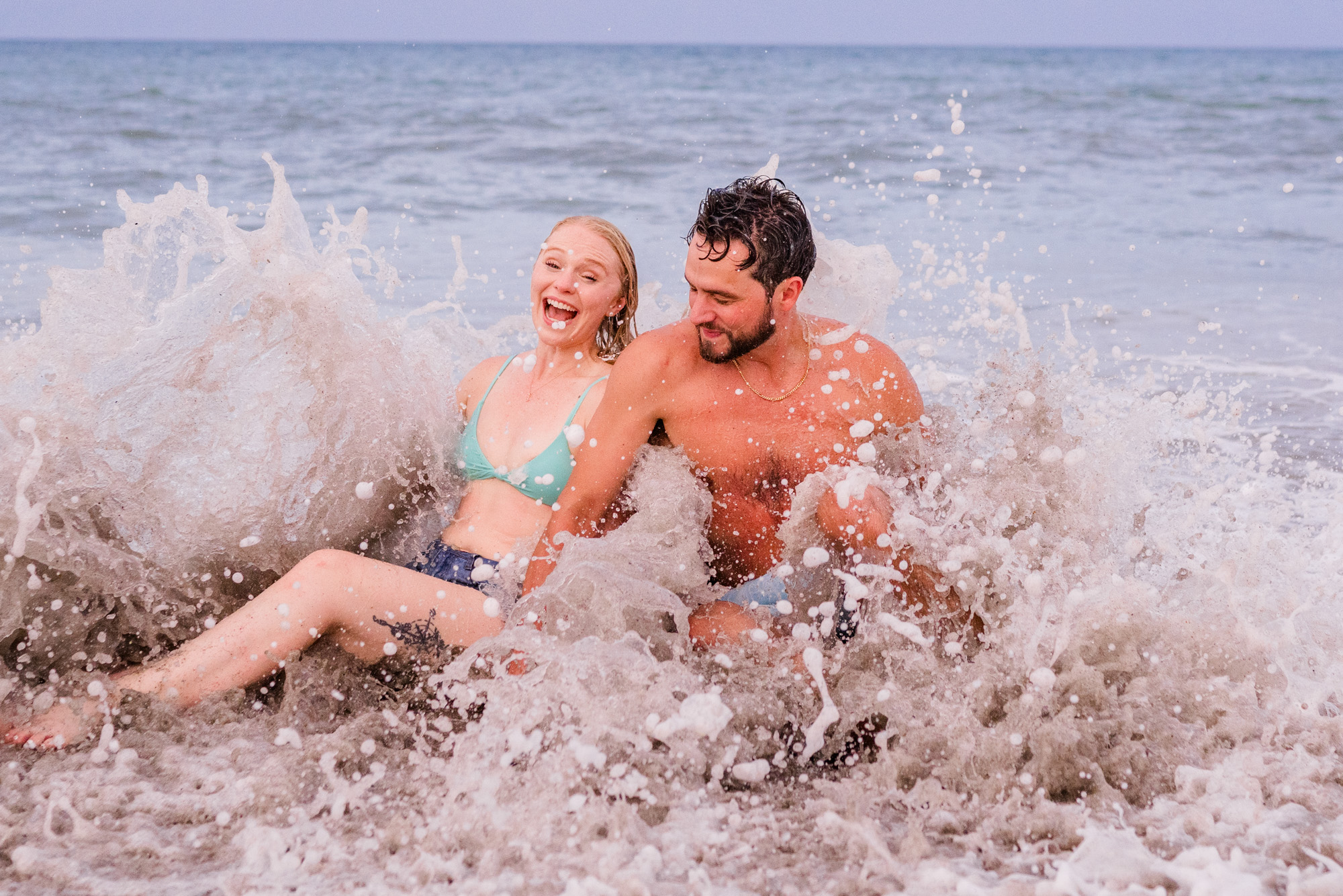 couple laughing while being splashed by a waved during their beach engagement session
