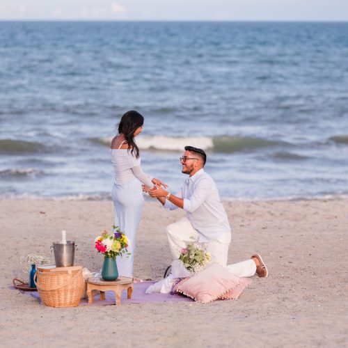 Man dressed in light blue and white down on one knee proposing to woman dressed in blue with the Atlantic Ocean behind them on Cocoa Beach, Florida