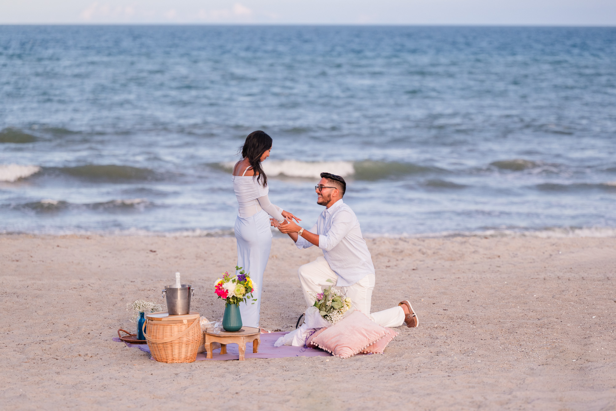 Man dressed in light blue and white down on one knee proposing to woman dressed in blue with the Atlantic Ocean behind them on Cocoa Beach, Florida