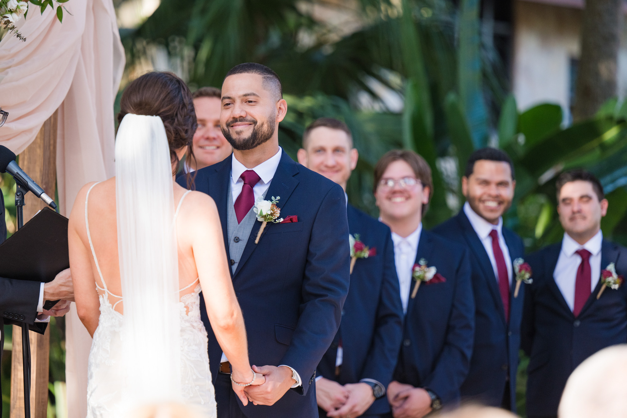 Groom smiling at the bride during their outdoor wedding ceremony with groomsmen behind him.