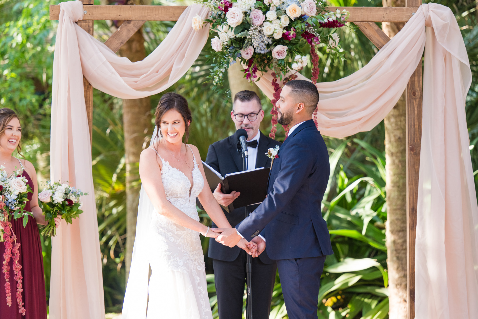 Bride and groom holding hands beneath a blush-draped floral arch during their wedding ceremony.