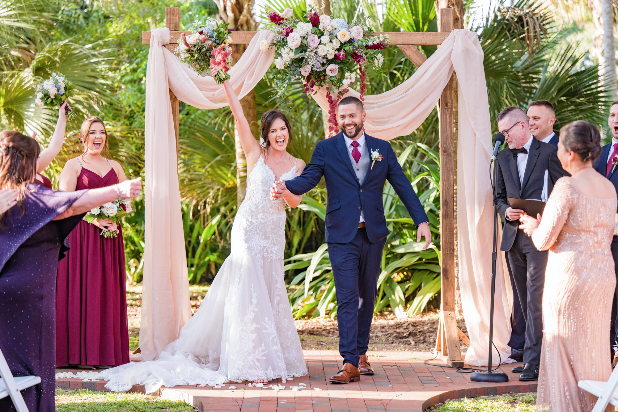 Newlyweds celebrating beneath the floral ceremony arch as the bride raises her bouquet in the air.