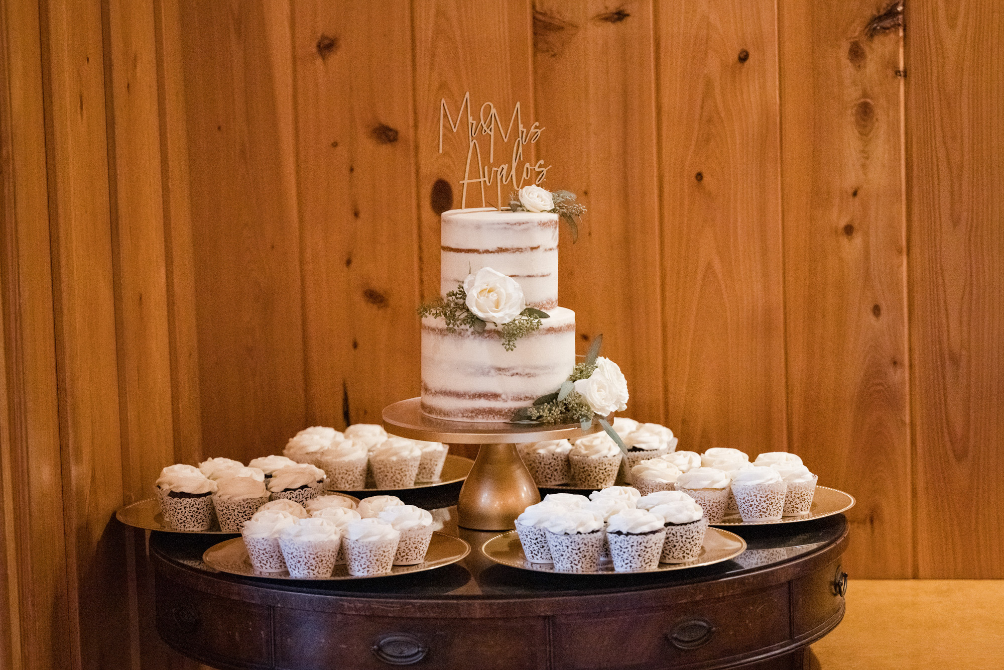 Two-tier wedding cake with white roses surrounded by cupcakes in the wood-paneled reception space.