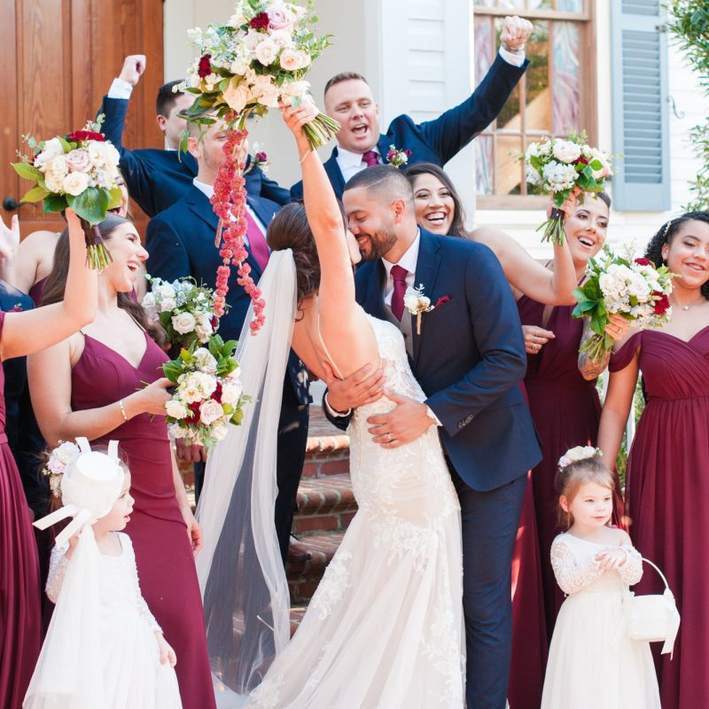 Bride raising her bouquet while the groom kisses her during a lively wedding party portrait.