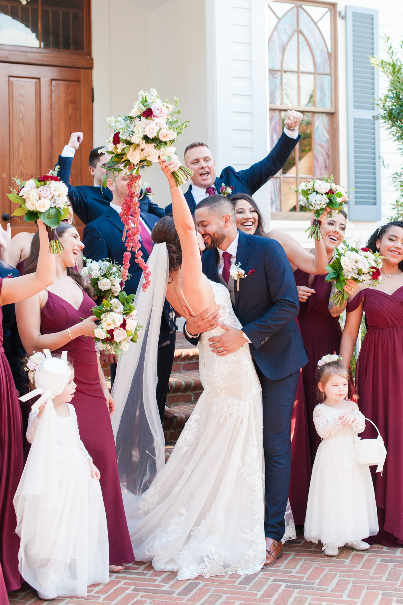 Bride raising her bouquet while the groom kisses her during a lively wedding party portrait.