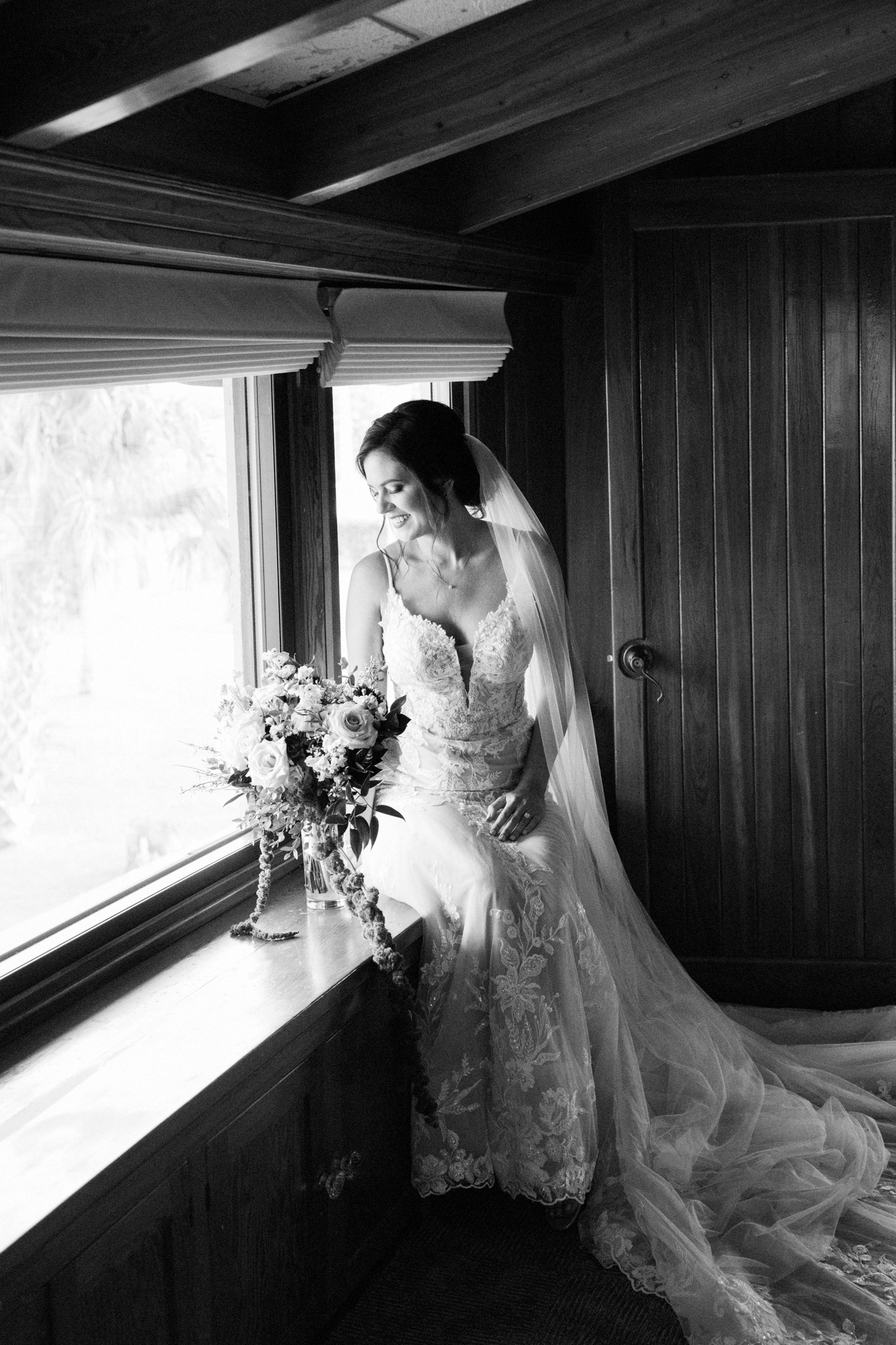 Bride sitting beside a window with her bouquet in a black-and-white wedding portrait.