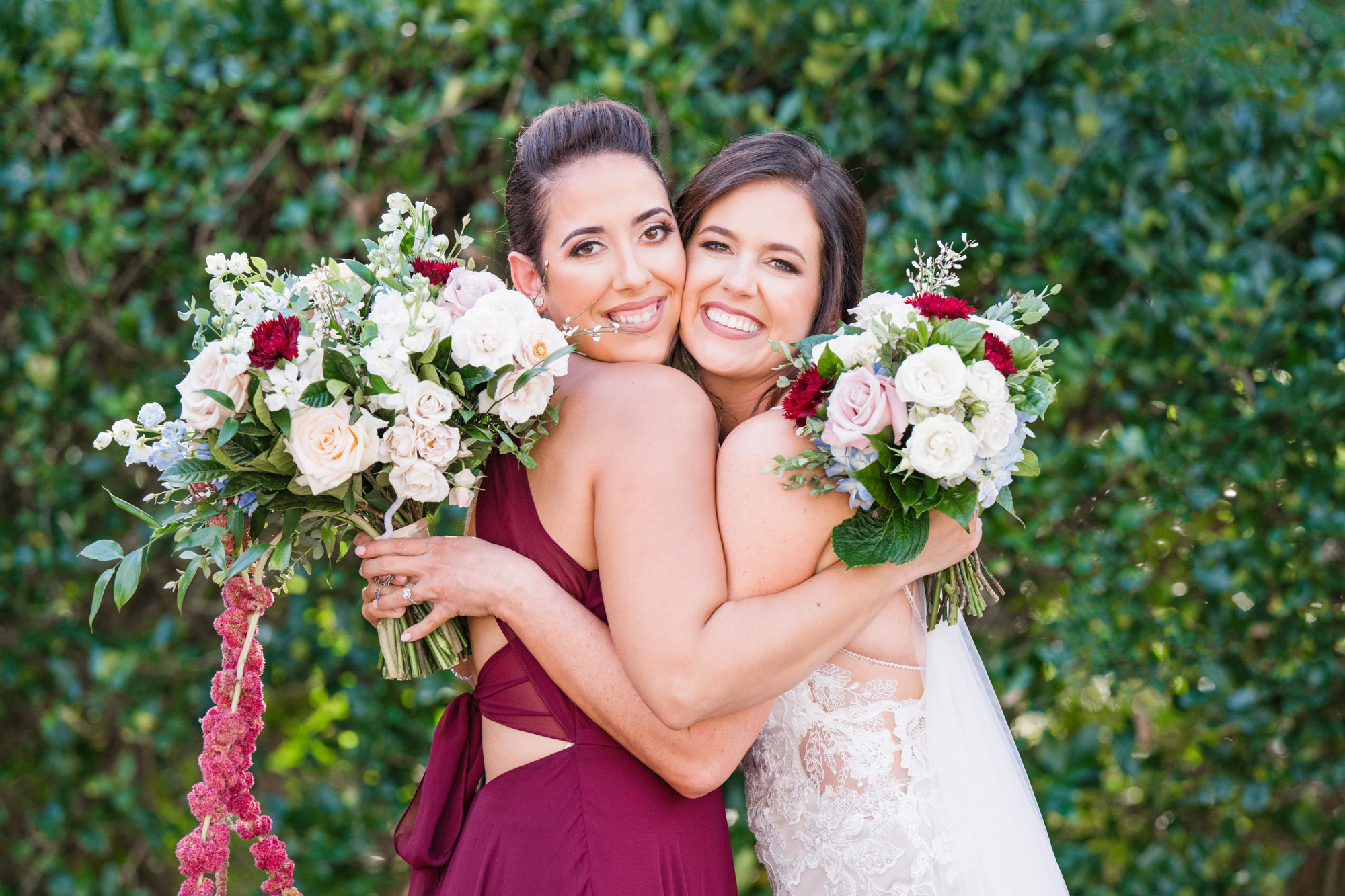 Bride hugging a bridesmaid while both hold floral bouquets during wedding portraits.