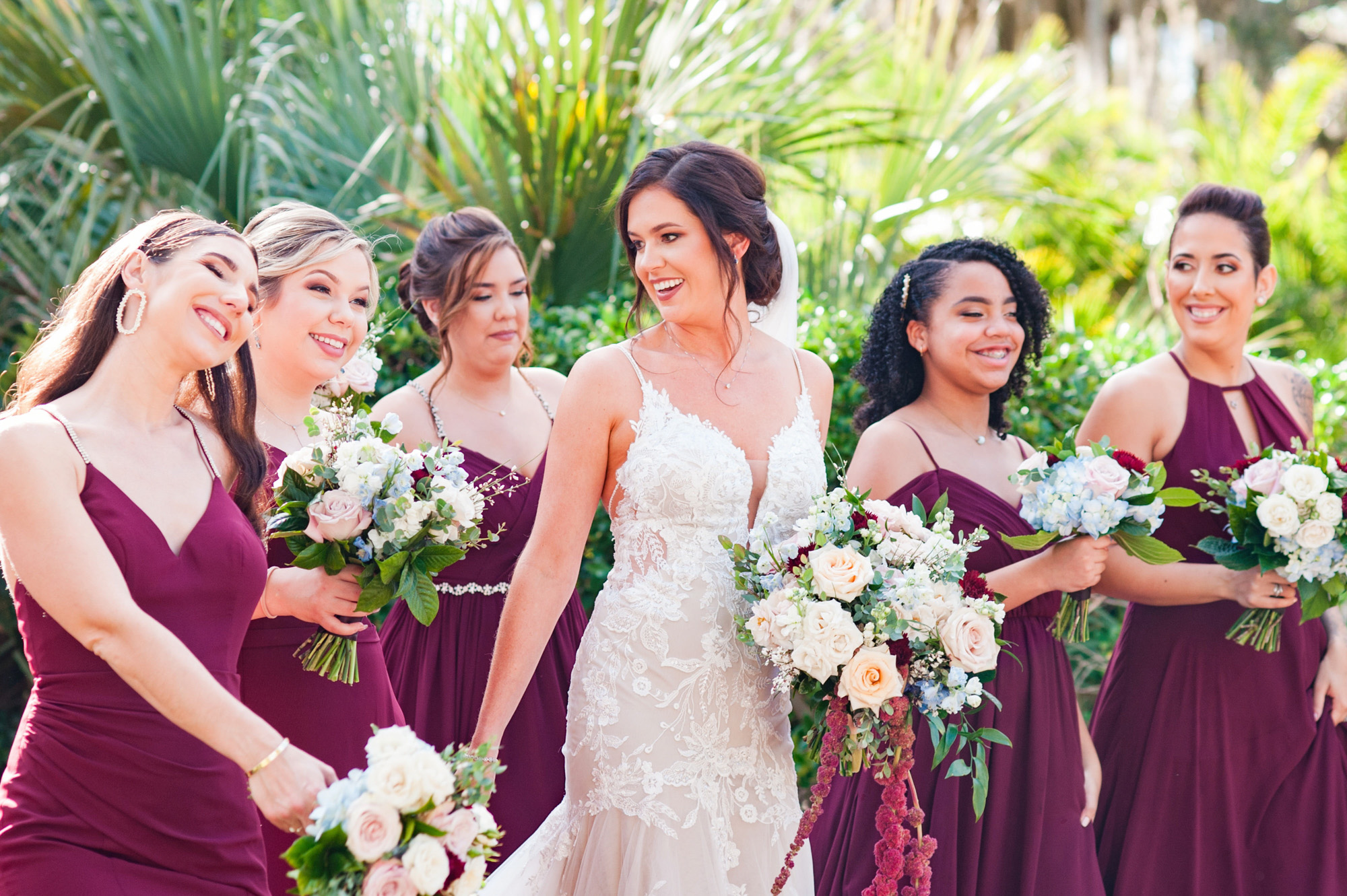 Bride laughing with her bridesmaids in burgundy dresses and carrying coordinated bouquets.