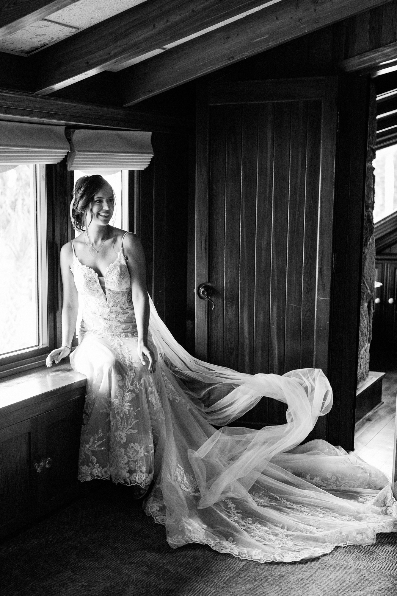 Black-and-white bridal portrait by a window with the bride’s long veil and lace train.
