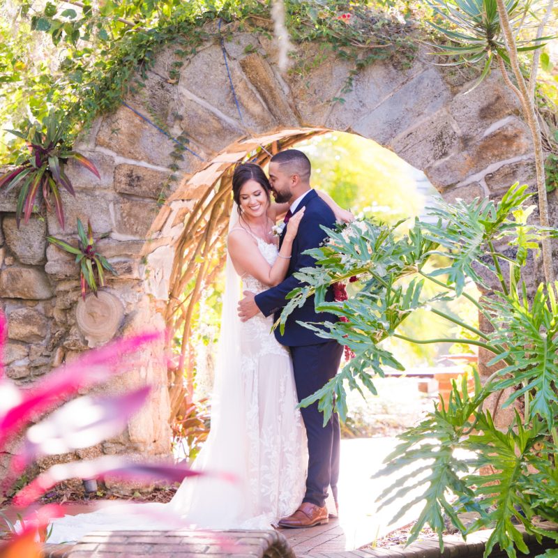 Bride and groom embracing beneath a stone garden arch surrounded by tropical greenery.