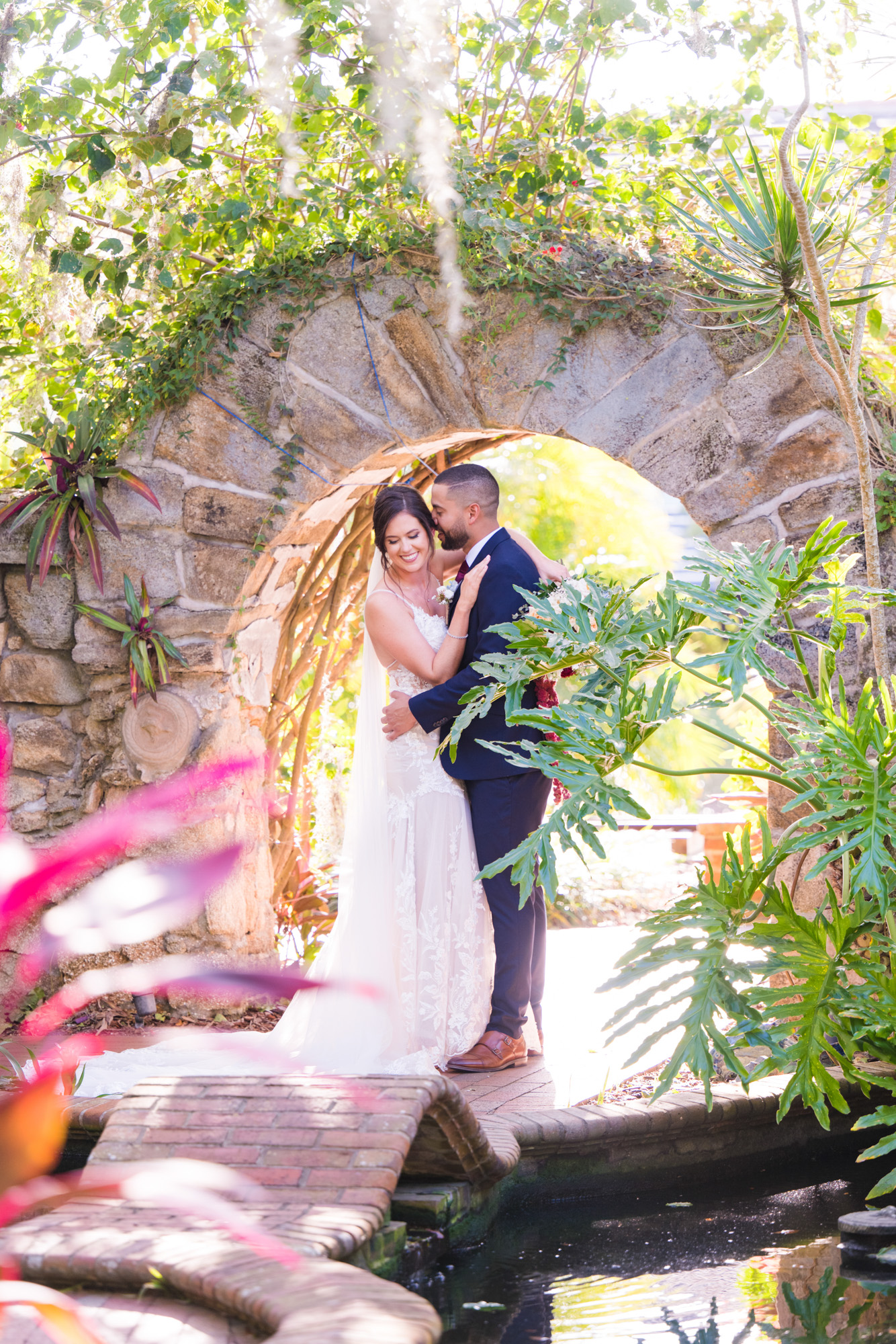 Bride and groom embracing beneath a stone garden arch surrounded by tropical greenery.