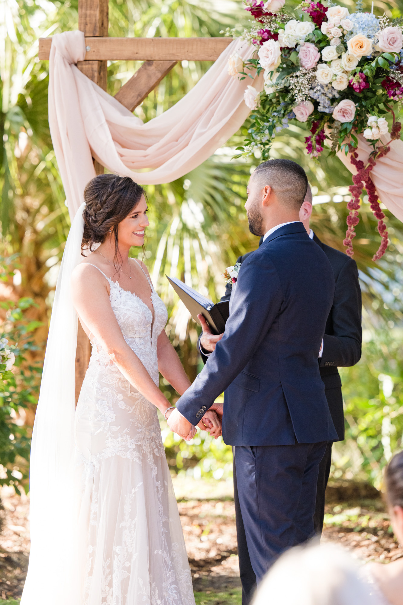 Bride and groom holding hands during their outdoor wedding ceremony beneath a floral arch.