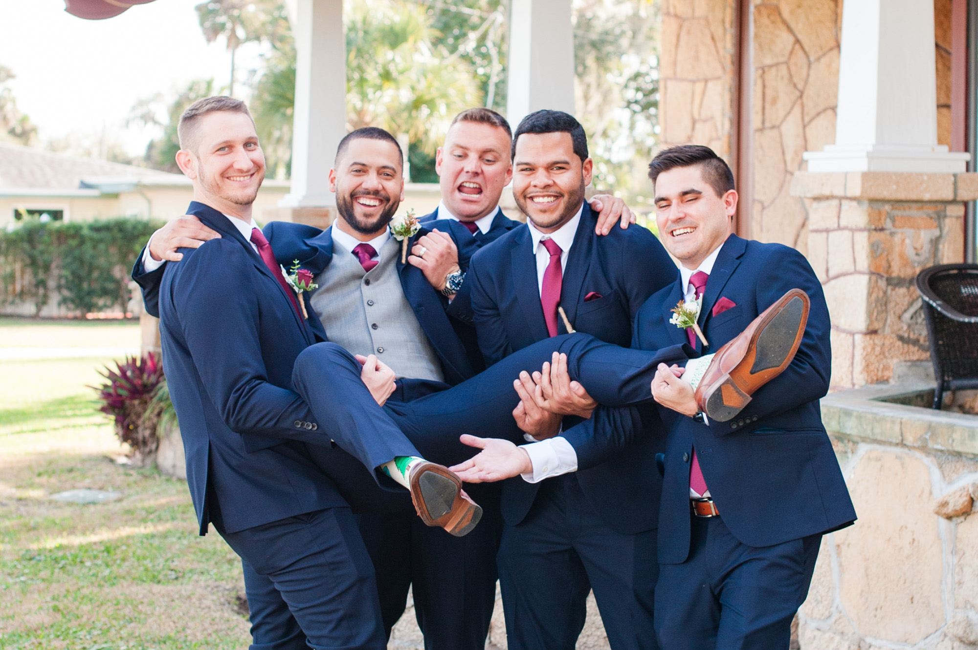 Groomsmen laughing while holding the groom during a playful wedding party portrait.