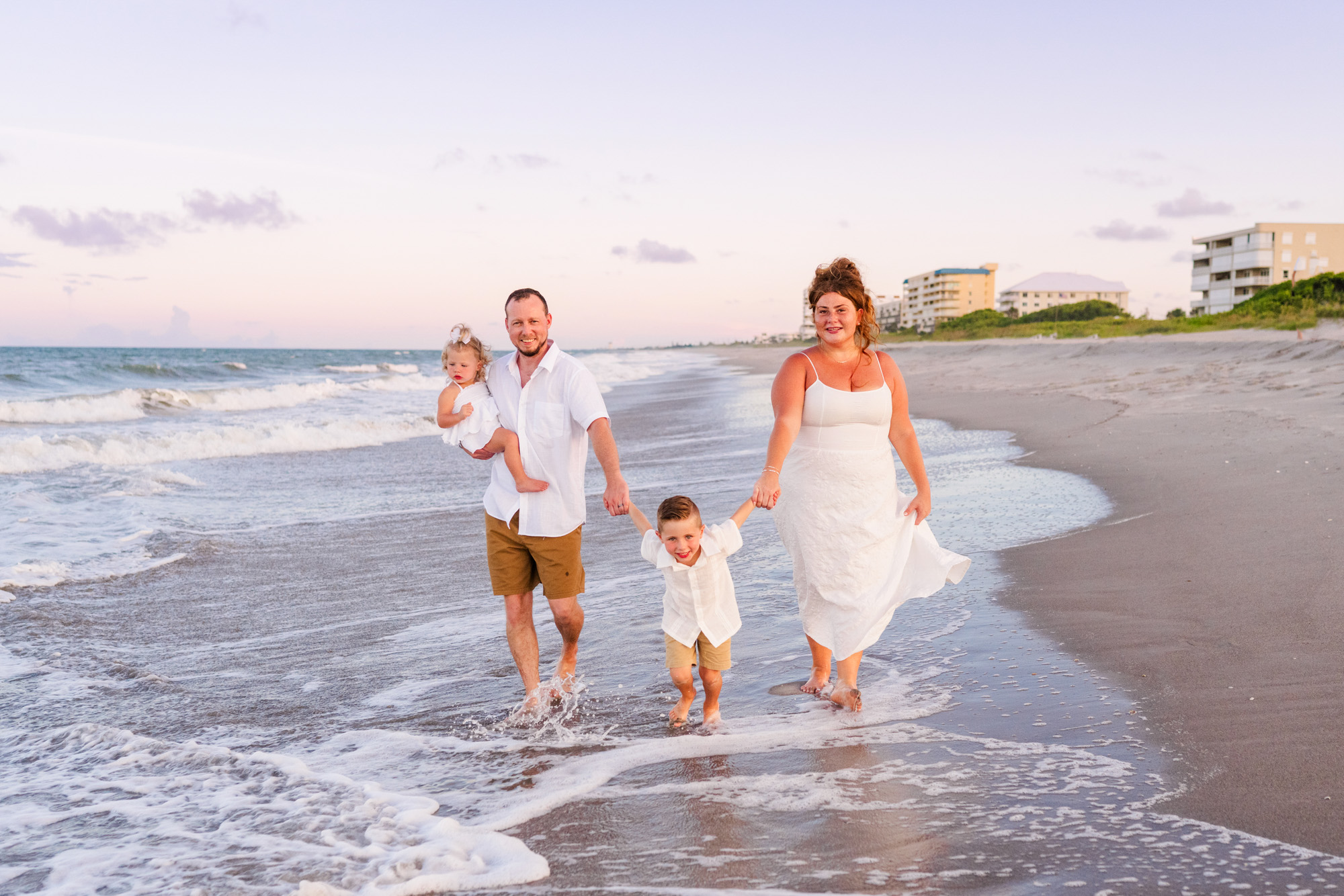 Family of four mostly dressed in white walking down the beach during their family session