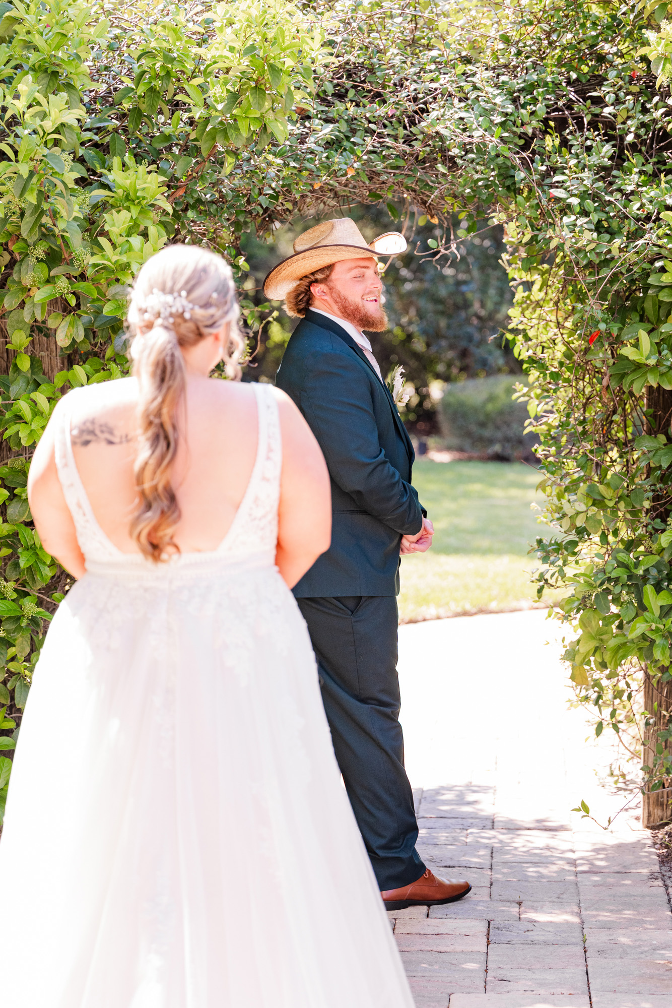 Bride approaches the groom in a cowboy hat for their wedding first look beneath a leafy garden arch.