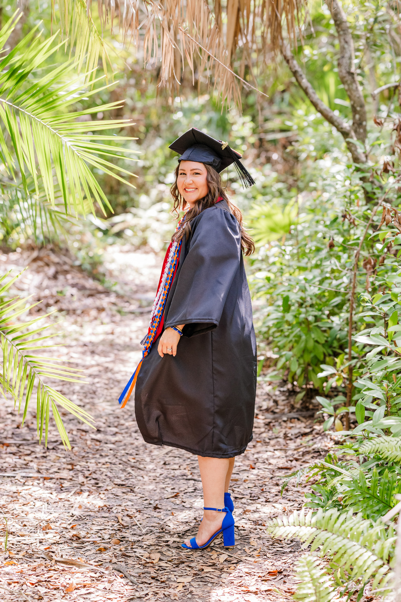Graduate posing in her cap and gown surrounded by palms and tropical greenery.