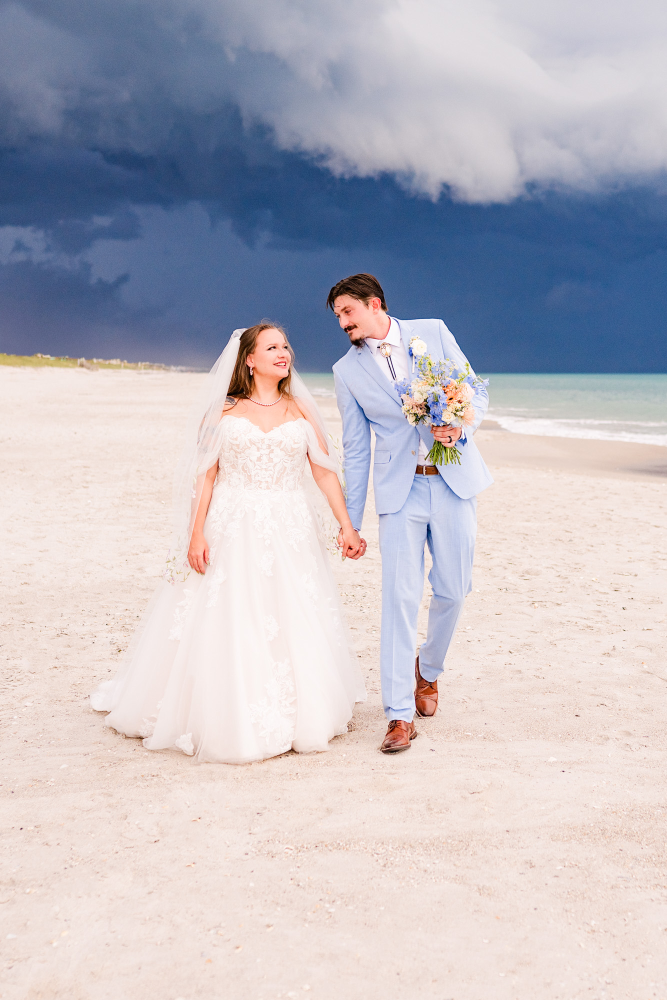 Bride and groom walking down the beach holding hands with a storm brewing