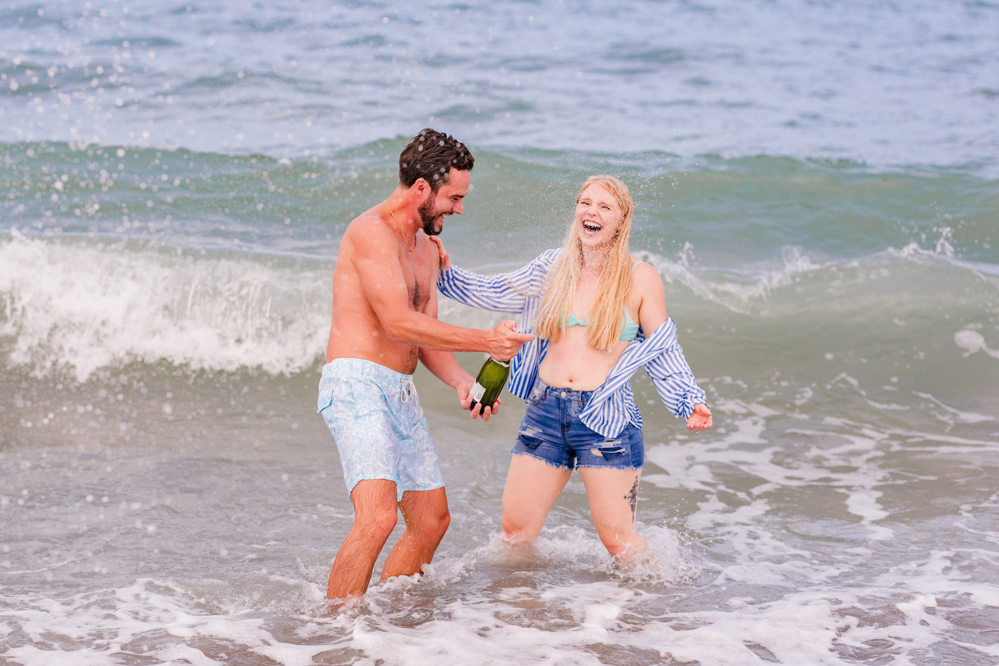 Couple popping champagne while playing in the ocean during their engagement session