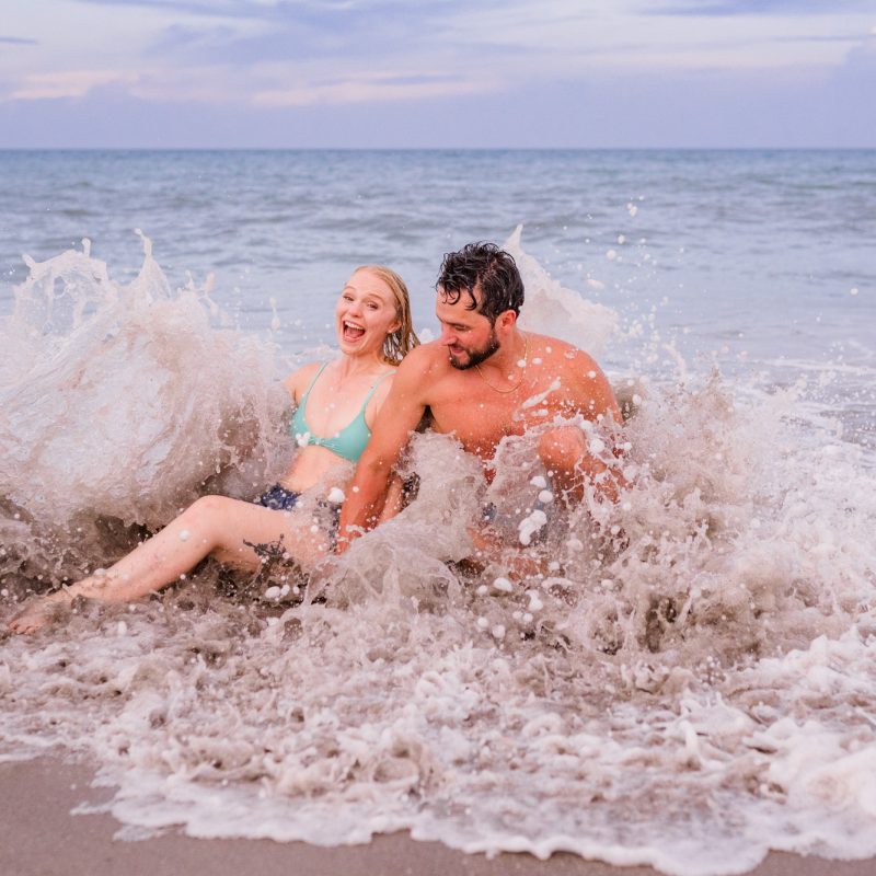 couple sitting in ocean with a giant wave splashing over them during their Melbourne Beach Engagement session