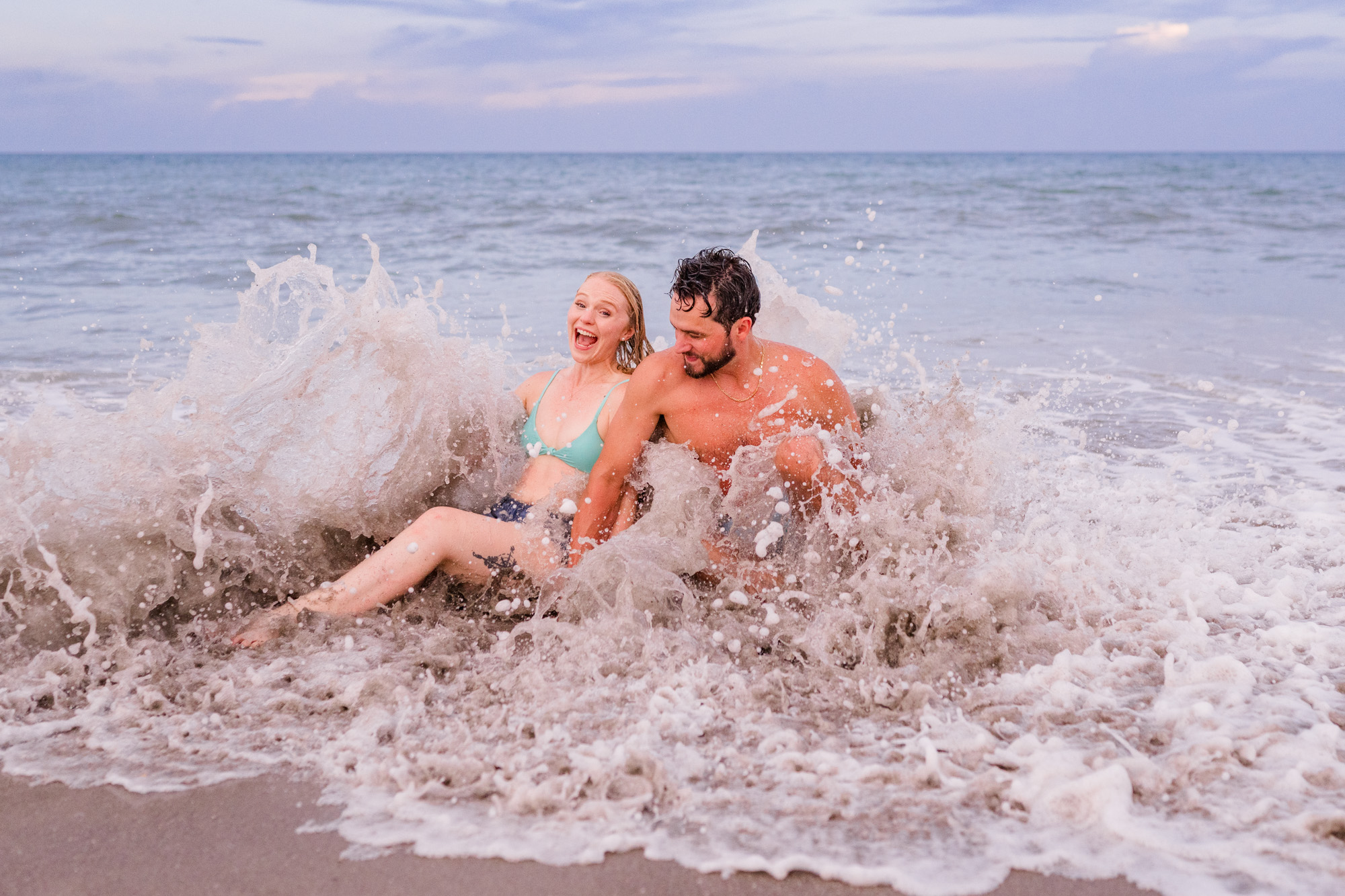 couple sitting in ocean with a giant wave splashing over them during their Melbourne Beach Engagement session