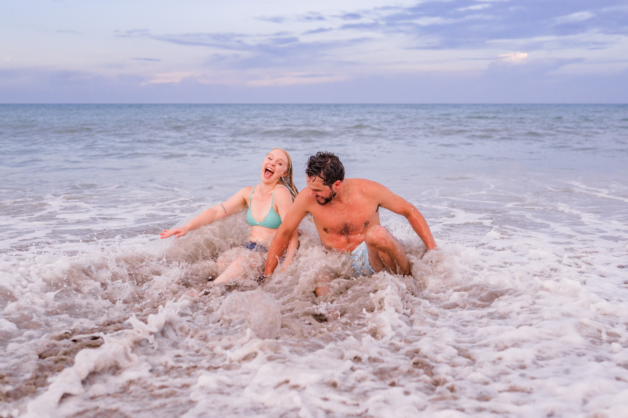 Couple sitting in the ocean with a wave crashing over them at their Melbourne Beach engagement session