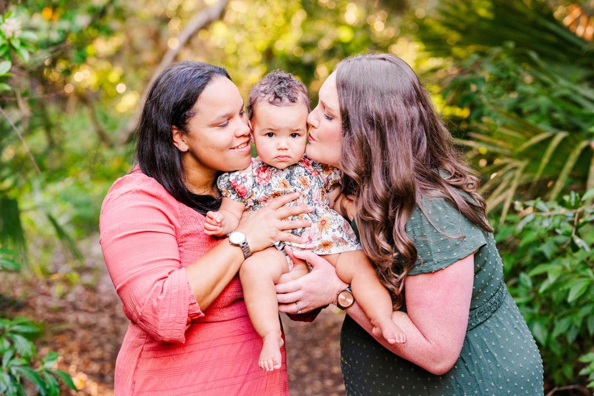two moms in the woods kissing their daughter during a melbourne beach family session