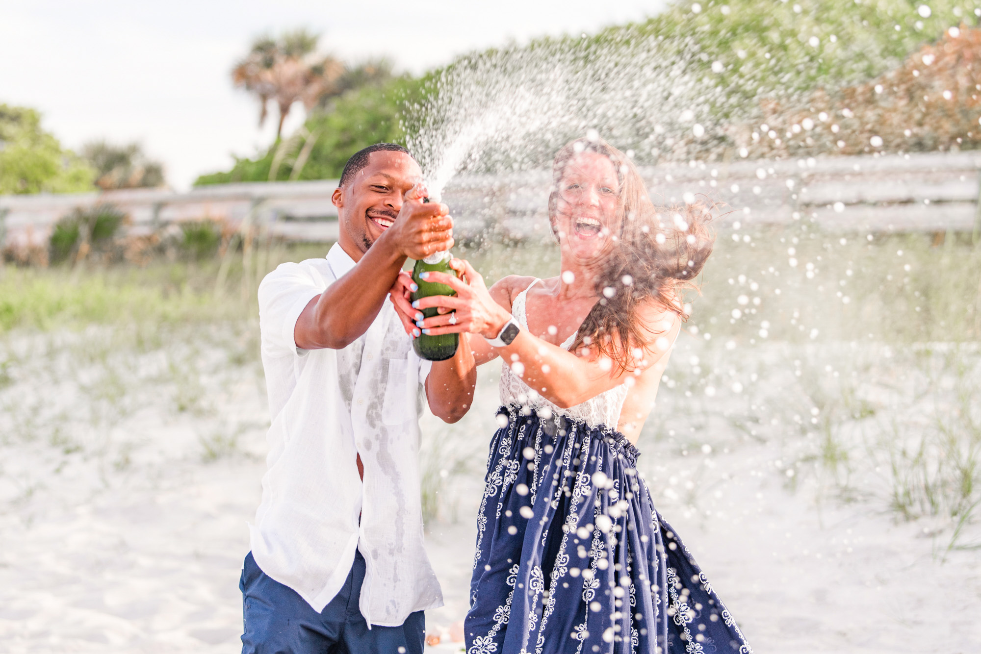 Couple popping champagne bottle on the beach after she said yes to marrying him