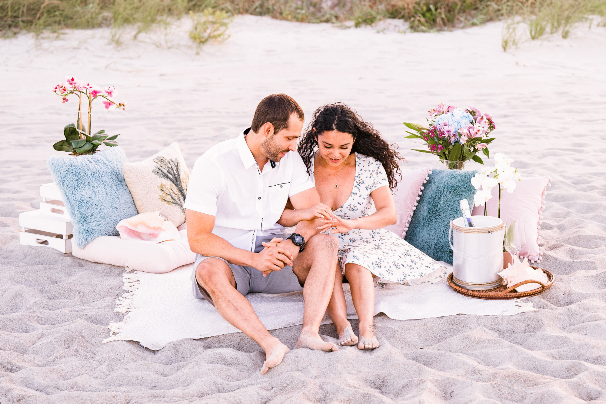 Man and woman on a beach blanket surrounded by flowers and pillows just after a marriage proposal