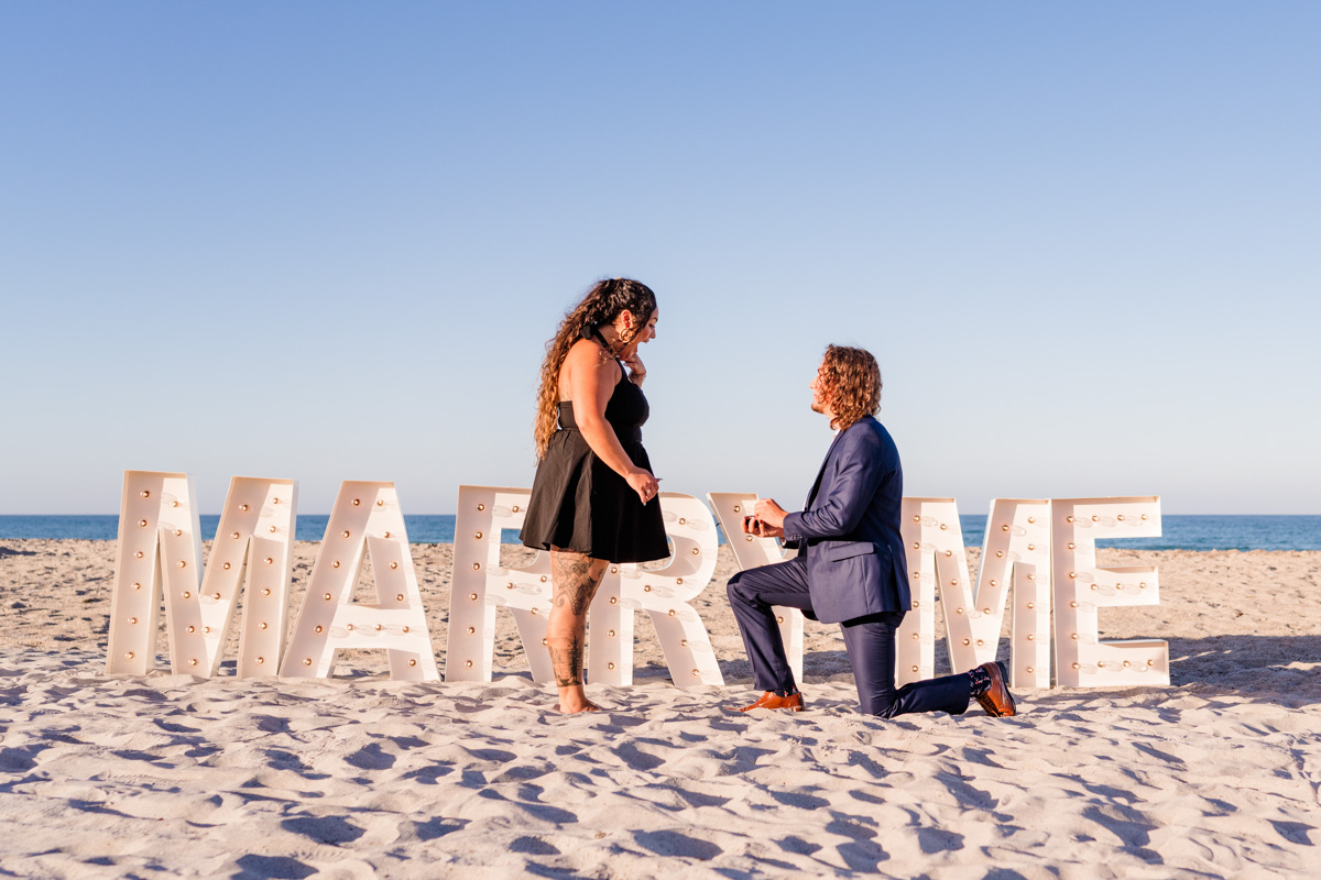 Man down on one knee in front of giant marquee letters saying marry me on Melbourne Beach