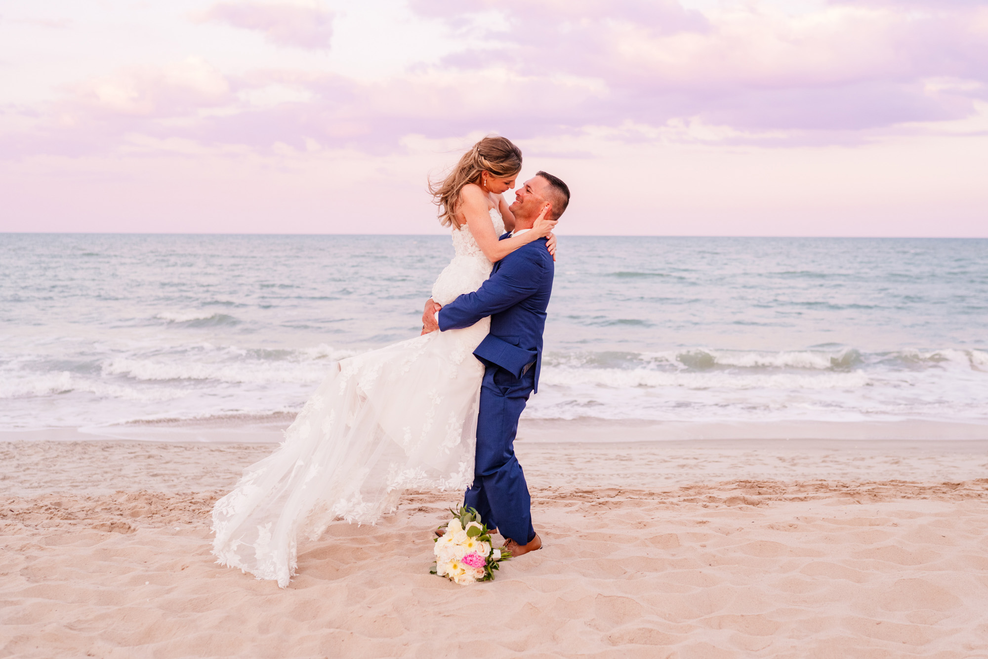 Groom lifting his bride in the air during a beautiful sunset with the ocean in the backgroun