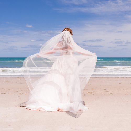 Bride facing the ocean with veil blowing in the wind on a sunny day on Melbourne Beach
